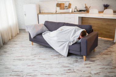 Grip olmuş genç bir adam, evdeki kanepede oturuyor, battaniyeye sarılmış..