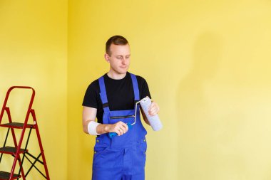 Tulumlu yakışıklı adam boya silindiri tutuyor. Apartman odası tadilatı için alet edevat. Ev yenileme konsepti