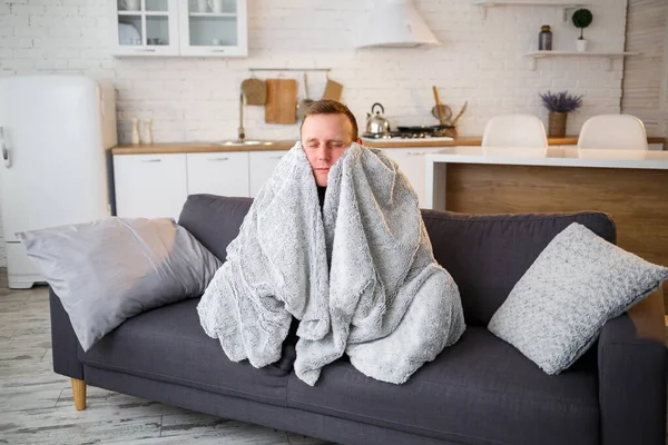 A sick man sits at home on a gray sofa with a white blanket. Illness, protection, coronavirus, illness, flu.