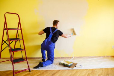 Bir adam evin duvarını silindir ve boyayla boyuyor. Evdeki odaların yenilenmesi.
