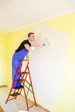 Bir adam evin duvarını silindir ve boyayla boyuyor. Evdeki odaların yenilenmesi.