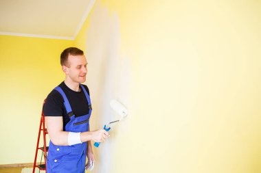 Tulumlu genç adam boya silindiri tutuyor. Apartman odası tadilatı için alet edevat. Ev yenileme konsepti