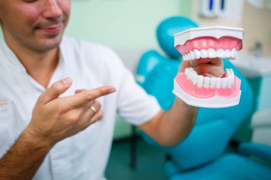 Beyaz üniformalı pozitif doktor dişçide otururken sahte çenesini dişleriyle gösteriyor.