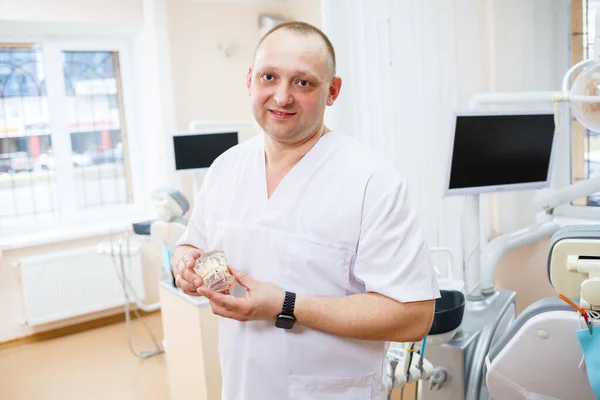 Portrait of smiling man in white doctor robe uniform is standing in office, clinic with chair, instruments. Dentist workplace. Orthodontist is preparing for visit of patient. Dentistry concept. — Stock Photo, Image