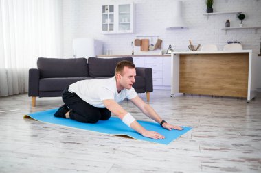 Genç adam evinde halıda oturuyor ve spor sonrası esneme hareketleri yapıyor.