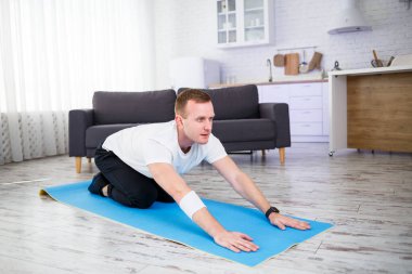 Genç adam evinde halıda oturuyor ve spor sonrası esneme hareketleri yapıyor.