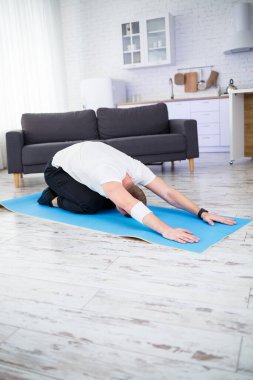 Genç adam evinde halıda oturuyor ve spor sonrası esneme hareketleri yapıyor.