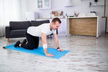 Genç adam evinde halıda oturuyor ve spor sonrası esneme hareketleri yapıyor.