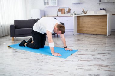 Genç adam evinde halıda oturuyor ve spor sonrası esneme hareketleri yapıyor.