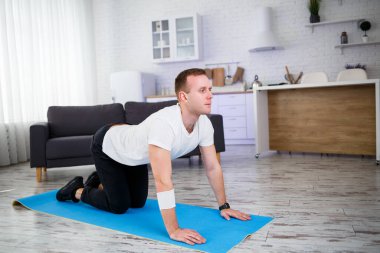 Genç adam evinde halıda oturuyor ve spor sonrası esneme hareketleri yapıyor.