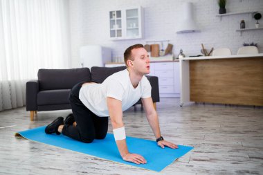 Genç adam evinde halıda oturuyor ve spor sonrası esneme hareketleri yapıyor.