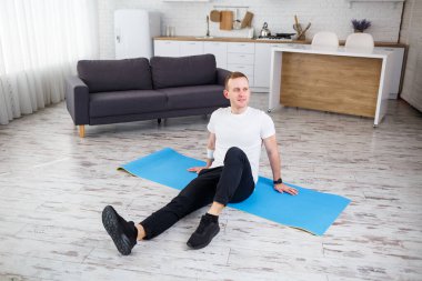 Genç adam evinde halıya oturmuş spor yaptıktan sonra dinleniyor.