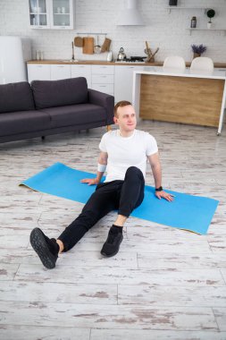 Genç adam evinde halıya oturmuş spor yaptıktan sonra dinleniyor.