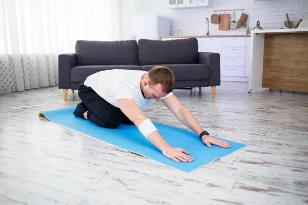 Young man sitting on carpet in his home and doing stretching after sports