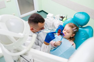 Profesyonel bir doktor, bir çocuk dişçisi, küçük bir kızın dişlerini aletlerle tedavi ediyor. Hasta muayenesi için dişçi ofisi. Bir çocukta diş tedavisi süreci