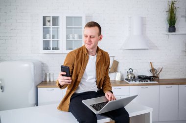 Gündelik giysiler içinde çekici bir genç adam mutfakta dizüstü bilgisayar kullanarak oturuyor. Evden çalışmak, uzak iş akışı.