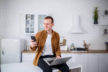 Gündelik giysiler içinde çekici bir genç adam mutfakta dizüstü bilgisayar kullanarak oturuyor. Evden çalışmak, uzak iş akışı.