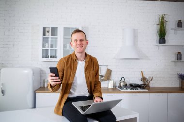 Gündelik giysiler içinde çekici bir genç adam mutfakta dizüstü bilgisayar kullanarak oturuyor. Evden çalışmak, uzak iş akışı.