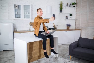 Gündelik giysiler içinde çekici bir genç adam mutfakta dizüstü bilgisayar kullanarak oturuyor. Evden çalışmak, uzak iş akışı.