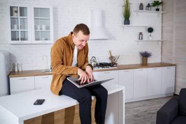 Gündelik giysiler içinde çekici bir genç adam mutfakta dizüstü bilgisayar kullanarak oturuyor. Evden çalışmak, uzak iş akışı.