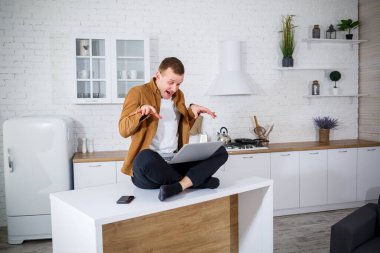 Gündelik giysiler içinde çekici bir genç adam mutfakta dizüstü bilgisayar kullanarak oturuyor. Evden çalışmak, uzak iş akışı.