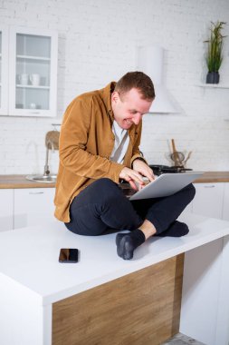Gündelik giysiler içinde çekici bir genç adam mutfakta dizüstü bilgisayar kullanarak oturuyor. Evden çalışmak, uzak iş akışı.