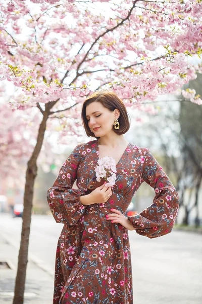 A spectacular woman in a bright dress stands against the background of sakura. A dark-haired woman in a beautiful outfit smiles on the street while walking