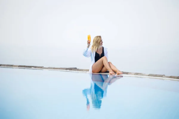 Una joven y hermosa mujer en traje de baño se sienta cerca de la piscina y unta su piel con un protector solar. Cuidado de la piel en el verano — Foto de Stock