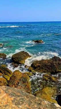 Sörfçüler, açık mavi gökyüzünün altında kayalık çıkıntılarda süzülürken nazik dalgalarda sörf yaparlar. Huzurlu sahne, doğanın ihtişamını ve rahatlığını davet eder.