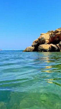 Deniz kenarında güneşli bir günde berrak bir turkuaz su, kayalık bir kıyı şeridi ve parlak bir gökyüzünü yansıtır. Huzurlu manzaraya doku ve güzellik katar.