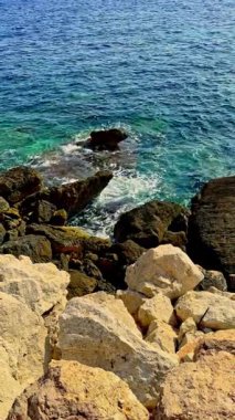 Sakin bir sahil manzarası, berrak mavi suların pürüzsüz kayalara çarptığını güneş ışığının yüzeyden yansıyarak doğal bir ortamda barışçıl bir atmosfer yarattığını gösteriyor.