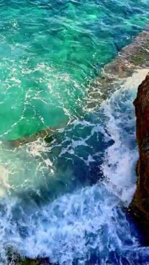 Dalgalar enerjik bir şekilde engebeli bir uçuruma doğru yuvarlanır ve parlak güneş ışığında beyaz köpük oluşturur. Berrak turkuaz su, kıyıdaki barışçıl yansımayı davet eder.