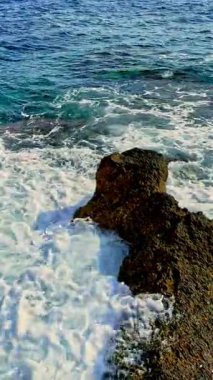 Köpüklü dalgalar parlak güneşin altındaki sivri kayaların etrafını sarar berrak mavi su parıldar kıyı şeridi boyunca huzur ve yansımayı davet eden huzurlu bir sahne yaratır.