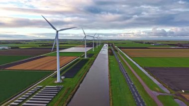 Rüzgâr türbinleri, yemyeşil tarlalar ve kahverengi tarım arazileriyle çevrili durgun bir kanalda, doğanın ahengi ve yenilenebilir enerjiyi yakalar.