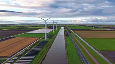 Geniş yeşil tarlalar kırsal alana yayılıyor. Uzun rüzgar türbinleri rüzgarda zarifçe dönüyor. Sakin bir su yolu, gökyüzünü yansıtıyor ve huzurlu manzarayı güzelleştiriyor.
