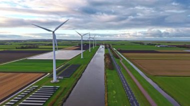 Uzun rüzgar türbinleri, canlı yeşil tarlalar ve doğa ve teknoloji arasındaki uyumu vurgulayan tarım arazileriyle çevrili dolambaçlı bir kanalın yanında gururla durur.
