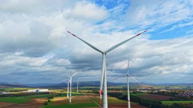 Büyük rüzgar türbinleri yeşil bir alanda, parlak mavi bir gökyüzünün altında, temiz enerjiyi ve kırsal alanda sürdürülebilirliği simgeleyen kabarık bulutlarla birlikte zarifçe döner.