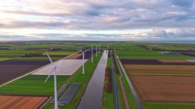 Rüzgar türbinleri, zengin yeşil alanlar ve sakin bir kanal ile çevrili, bulutlu bir gökyüzü altında doğa ve teknolojinin mükemmel bir karışımını sunan pitoresk bir arazide dimdik dururlar.