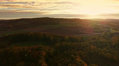 Güneş doğarken tepeleri kaplayan yumuşak ışık, sıcak sonbahar renklerinde boyanmış tarım arazileri ve ağaçlar kırsal alanda sakin bir sabah atmosferi yaratıyor.