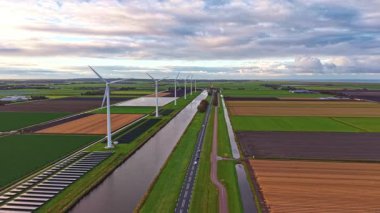 Rüzgâr türbinleri, yemyeşil tarlalarla çevrili bir kanal boyunca dimdik ayakta duruyor. Bu dingin ortamda akşam alacakaranlığında bulutlar yukarıda toplanıyor.