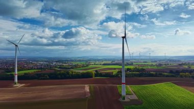 Uzun rüzgâr türbinleri yemyeşil tarlaların ve kahverengi yamaların üzerinde yükseliyor. Arkaplandaki bulutlar ve yuvarlanan tepeler, sakin bir kırsal bölgede yenilenebilir enerji anlarını yakalıyor.