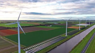 Rüzgâr türbinleri, parlak bir tarım sahnesinde, ufuk boyunca uzanan yeşil tarlaları ve zengin toprak renkleriyle güzel bir günbatımı gökyüzünün altında dimdik duruyorlar.