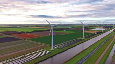 Rüzgar türbinleri alacakaranlıkta manzaralı bir kırsal alanda yavaşça döner yemyeşil tarlaların üzerine gölgeler ve sürdürülebilir enerjiyi ve doğayı sembolize eden renkli ekin sıraları serer.