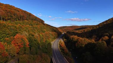 Sonbahar yapraklarıyla bezenmiş dağların arasından geniş bir yol rüzgarı. Güneş ışığı huzurlu kırsal bölgeye sıcak bir parıltı saçar ve sakin bir yolculuğa davet eder.