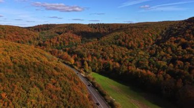 Yumuşak bulutlarla dolu mavi gökyüzünün altında dolambaçlı bir yol, huzurlu bir kırsal araziyi keserken, hareketli sonbahar yaprakları yuvarlanan tepeleri kaplıyor.