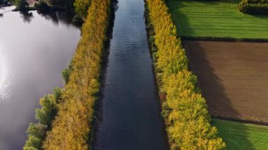 Altın yapraklar kıvrımlı bir nehrin her iki tarafında da uzanır. Temiz bir gökyüzü altında yeşil alanlar ise sonbaharda kırsal ortamın sakinliğini gösterir.