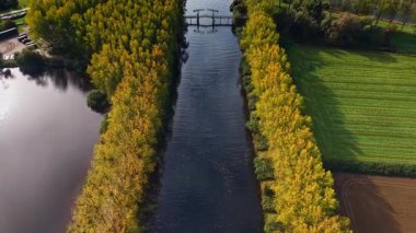 Parlak sarı ve yeşil ağaçlar, berrak gökyüzünün altındaki sonbahar renklerini yansıtan huzurlu nehir boyunca uzanan tarlalar boyunca suları aşan büyüleyici bir ahşap köprü.