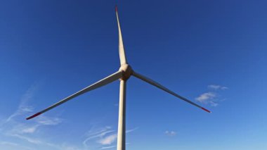Rüzgar enerjisini dizginlemeye hazır büyük bir rüzgar türbini gökyüzüne doğru uzanır. Sahne parlak ve güneşlidir. Sürdürülebilir bir gelecek için umut ve ilerleme hissi yaratır.