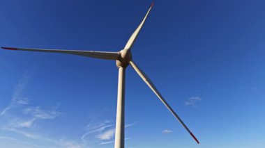 Sürdürülebilir yaşam için temiz enerji üretirken zarif bir şekilde dönen parlak mavi gökyüzünün arka planına karşı büyük bir rüzgar türbini yükselir.