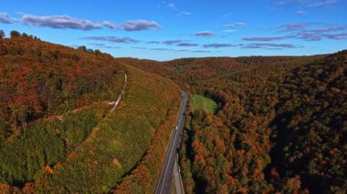 Parlak turuncu ve sarı yapraklar göz kamaştırıcı bir sonbahar sahnesi yaratır. Tepeler boyunca parlak mavi bir gökyüzü altında tüylü bulutlarla kaplı bir yol rüzgarı gibi.
